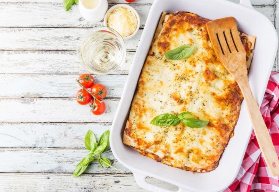 Freshly baked lasagna in dish with basil, cherry tomatoes, cheese, wine, and checkered cloth.