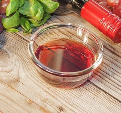 Glass bowl with red liquid, basil, grapes, and bottle with red cap on wooden surface.