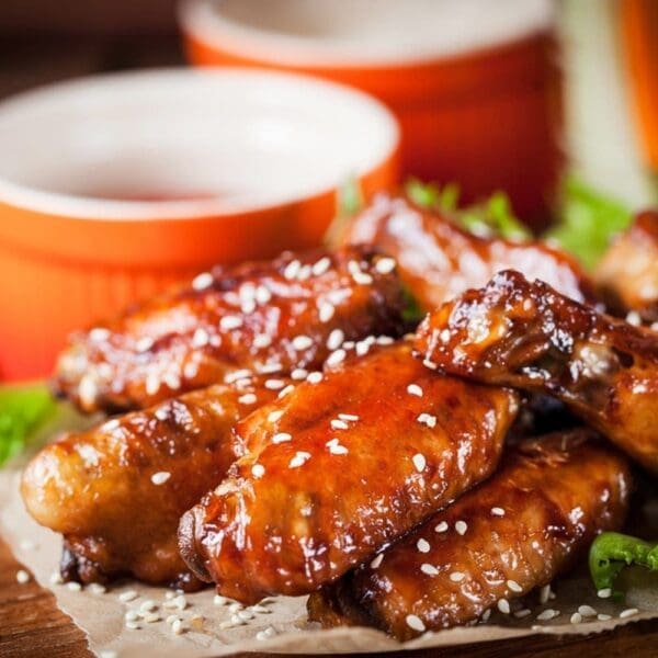 Plate of glazed chicken wings with sesame seeds on parchment, orange bowls in background.