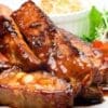 Glazed meat ribs on a plate with salad, sliced tomatoes, and coleslaw in the background.