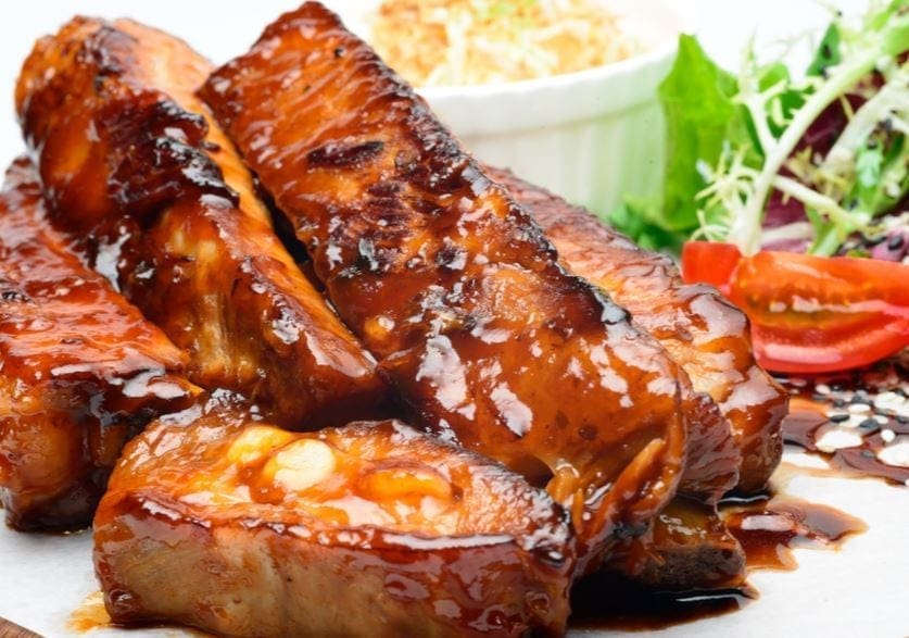 Glazed meat ribs on a plate with salad, sliced tomatoes, and coleslaw in the background.