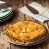Golden-brown pie with flaky crust and "I" cut, herbs on edge, plates, and napkin nearby.