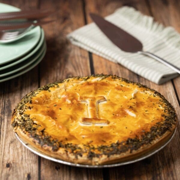 Golden-brown pie with flaky crust and "I" cut, herbs on edge, plates, and napkin nearby.