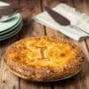 Golden-brown pie with sesame crust and embossed "I" on wooden table, plates, utensils nearby.