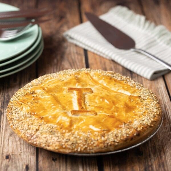 Golden-brown pie on wood surface with shiny crust, sesame edges, plates, knife, napkin nearby.