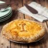 Golden pie with "I" on top, sesame seeds, green plates, striped napkin, and pie server nearby.