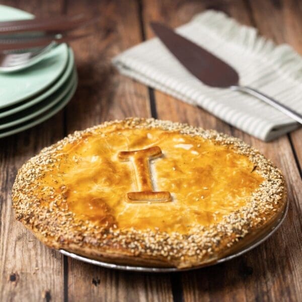 Golden pie with "I" on top, sesame seeds, green plates, striped napkin, and pie server nearby.