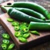 Green chili peppers, whole and sliced, on a wooden cutting board with visible seeds.