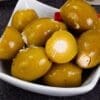 White dish with green olives and red pepper pieces. Some olives are stuffed with white filling.