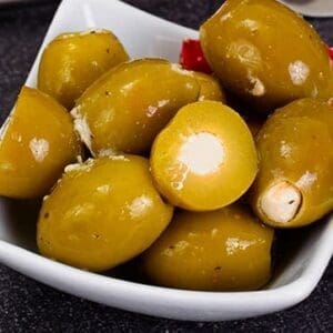 White dish with green olives and red pepper pieces. Some olives are stuffed with white filling.