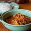 Green bowl of stew with meat, vegetables on wooden table; plate of white rice beside.
