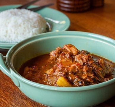 Green bowl of stew with meat, vegetables on wooden table; plate of white rice beside.