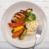 Grilled chicken with sauce, roasted vegetables, rice, and parsley on a white plate with fork.