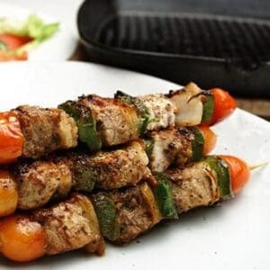 Grilled meat skewers with bell peppers, tomatoes, and onions on a plate near a grill pan.