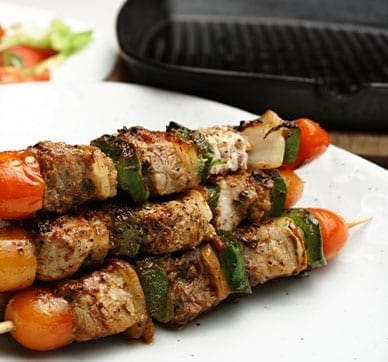 Grilled meat skewers with bell peppers, tomatoes, and onions on a plate near a grill pan.