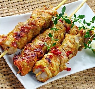 Grilled meat skewers with herbs and red pepper flakes on a white plate, wicker placemat background.