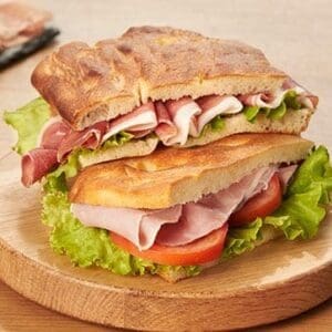Sandwich with ham, lettuce, and tomato on toasted bread on a round wooden serving board.