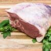 Large raw meat with fat on a wooden board, garnished with green herbs beneath.