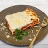 Plate of lasagna with layers, cheese, parsley, and golden fork on wooden background.
