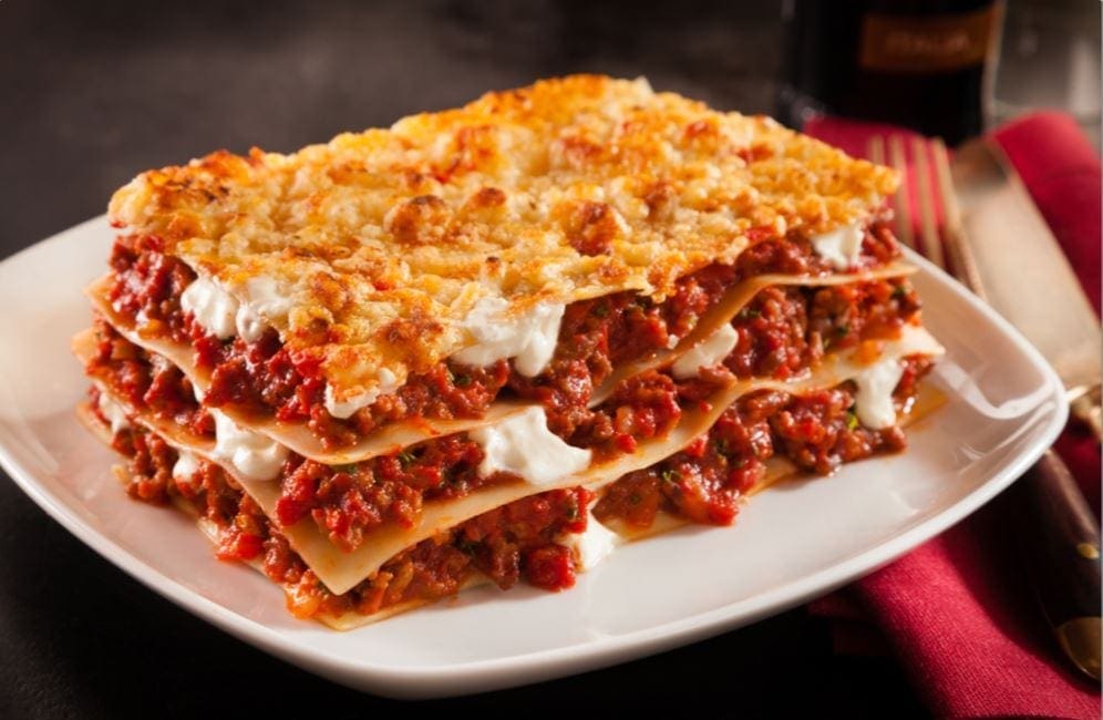 A plate of lasagna with layered pasta, meat sauce, cheese, on white plate, red napkin nearby.