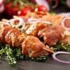 Marinated meat skewers with fresh herbs, sliced red onions, and blurred cherry tomatoes.
