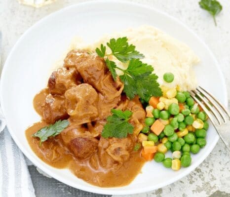 Meat in brown sauce with herbs, mashed potatoes, mixed vegetables, fork on plate.