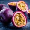 Passion fruits on dark surface, some whole with purple skins, others halved showing yellow interiors.