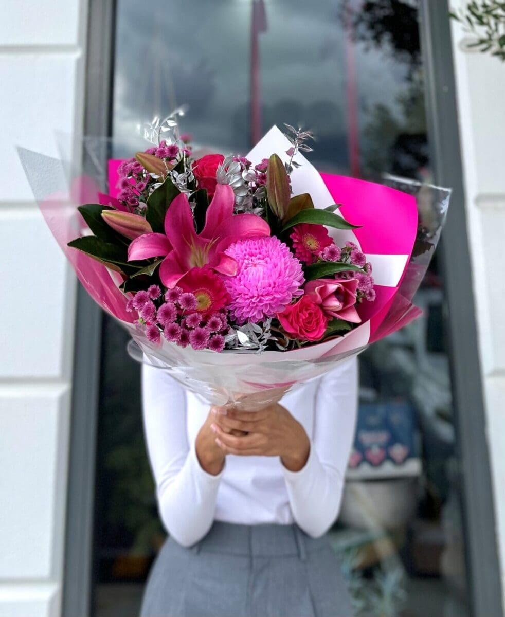 Person in white shirt holding pink flower bouquet with lilies and roses, building in background.