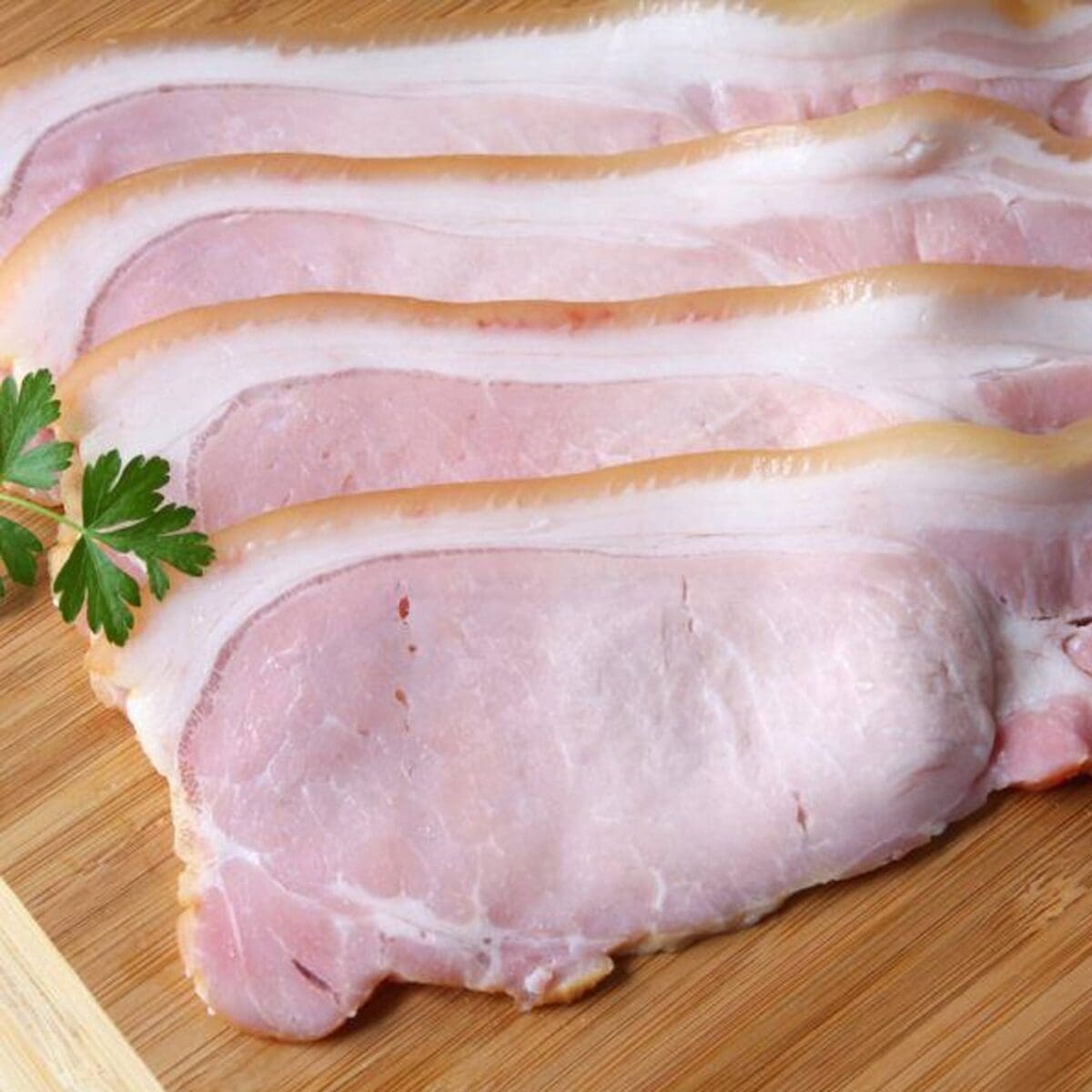 Raw bacon slices on a wooden board with a parsley sprig on the side.