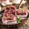 Raw beef medallions with bacon and rosemary in a bowl on wooden table.
