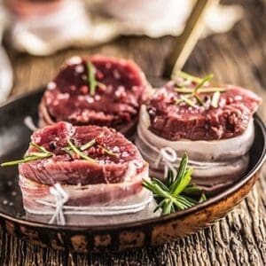 Raw beef medallions with bacon and rosemary in a bowl on wooden table.