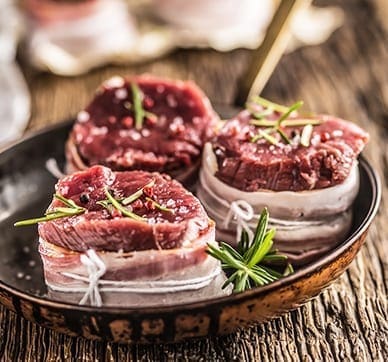 Raw beef medallions with bacon and rosemary in a bowl on wooden table.