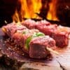 Raw beef skewers with bell peppers, coarse salt, and flames on a wooden board.
