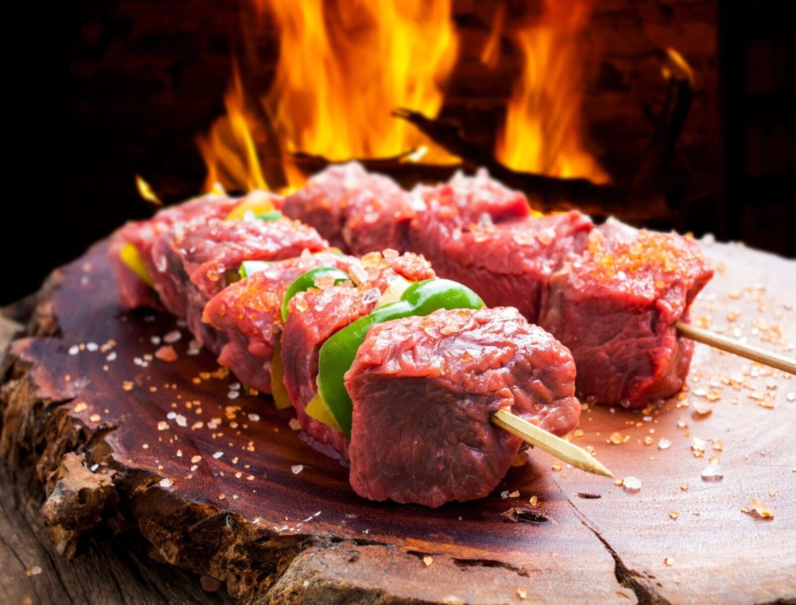 Raw beef skewers with bell peppers, coarse salt, and flames on a wooden board.