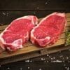 Raw beef steaks on wooden board with coarse salt and fresh herbs.