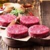 Raw burger patties on parchment paper with rosemary and chili peppers on a wooden board.