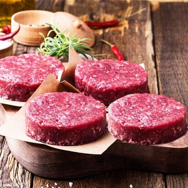 Raw burger patties on parchment paper with rosemary and chili peppers on a wooden board.