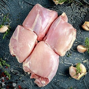 Four raw chicken breasts with garlic cloves and herbs on a dark surface.