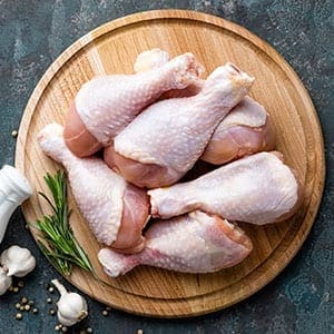 Raw chicken drumsticks on a wooden platter with garlic, peppercorns, and rosemary nearby.
