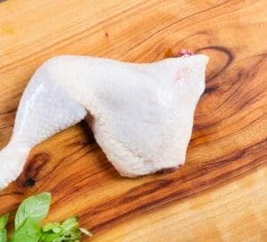 Raw chicken leg quarter on a wooden cutting board with fresh herbs.