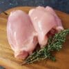 Two raw chicken thighs on a wooden board with a sprig of fresh herbs.