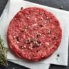 Raw hamburger patty on parchment with salt, pepper, herbs, and thyme on dark slate surface.