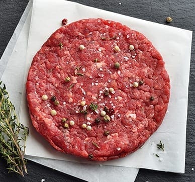 Raw hamburger patty on parchment with salt, pepper, herbs, and thyme on dark slate surface.