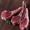 Three raw lamb chops on a wooden surface with a sprig of fresh mint.