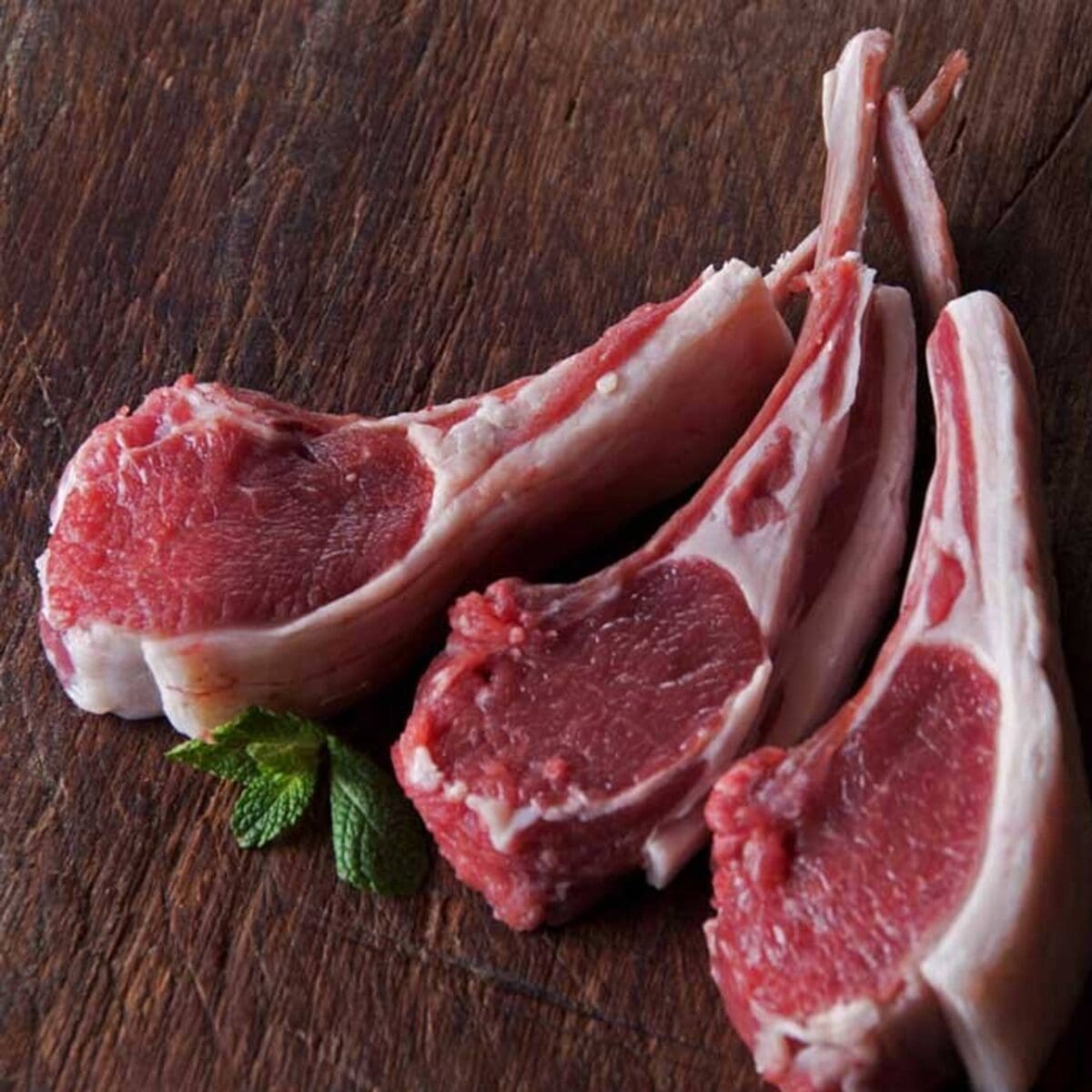 Three raw lamb chops on a wooden surface with a sprig of fresh mint.
