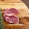 Two raw lamb chops on wooden board with fresh rosemary sprigs.