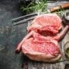 Raw marbled pork chops on wood with rosemary, fork, seasoning, and glass jar visible.