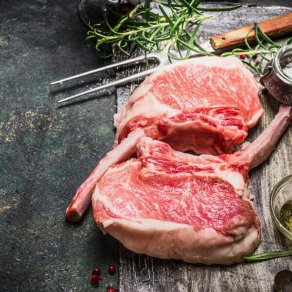 Raw marbled pork chops on wood with rosemary, fork, seasoning, and glass jar visible.