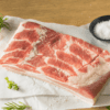 Raw pork belly on parchment, black dish with salt, herbs on wooden surface.