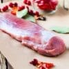 Raw pork tenderloin on brown paper with cranberries, bay leaves, and red peppers nearby.